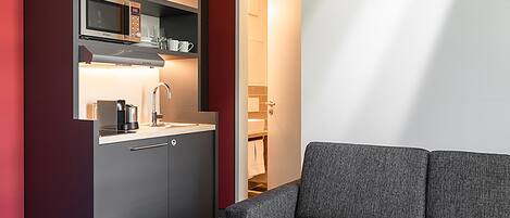 A modern kitchen with a microwave, coffee maker, and cabinets. There is a gray sofa and a small table with a white mug.