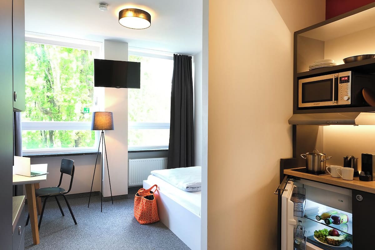 A hotel room with a bed, a desk with a laptop, a chair, a floor lamp, a window with a view of trees, a microwave, a refrigerator with food and drinks, and a shelf with plates and a bowl.