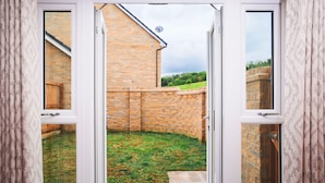 A view through a glass door into a backyard with a brick wall, a wooden fence, and a grassy area. - Peak District New Modern Cottage in Buxton (Buxton)