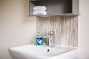 A bathroom sink with a bottle of hand soap, a towel shelf above it, and a wooden panel wall. - Peak District New Modern Cottage in Buxton (Buxton)