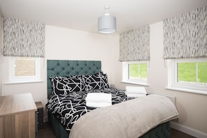 A bedroom with a tufted headboard, a bed with patterned bedding, two windows with blinds, a bedside table, and a hanging light fixture. - Peak District New Modern Cottage in Buxton (Buxton)