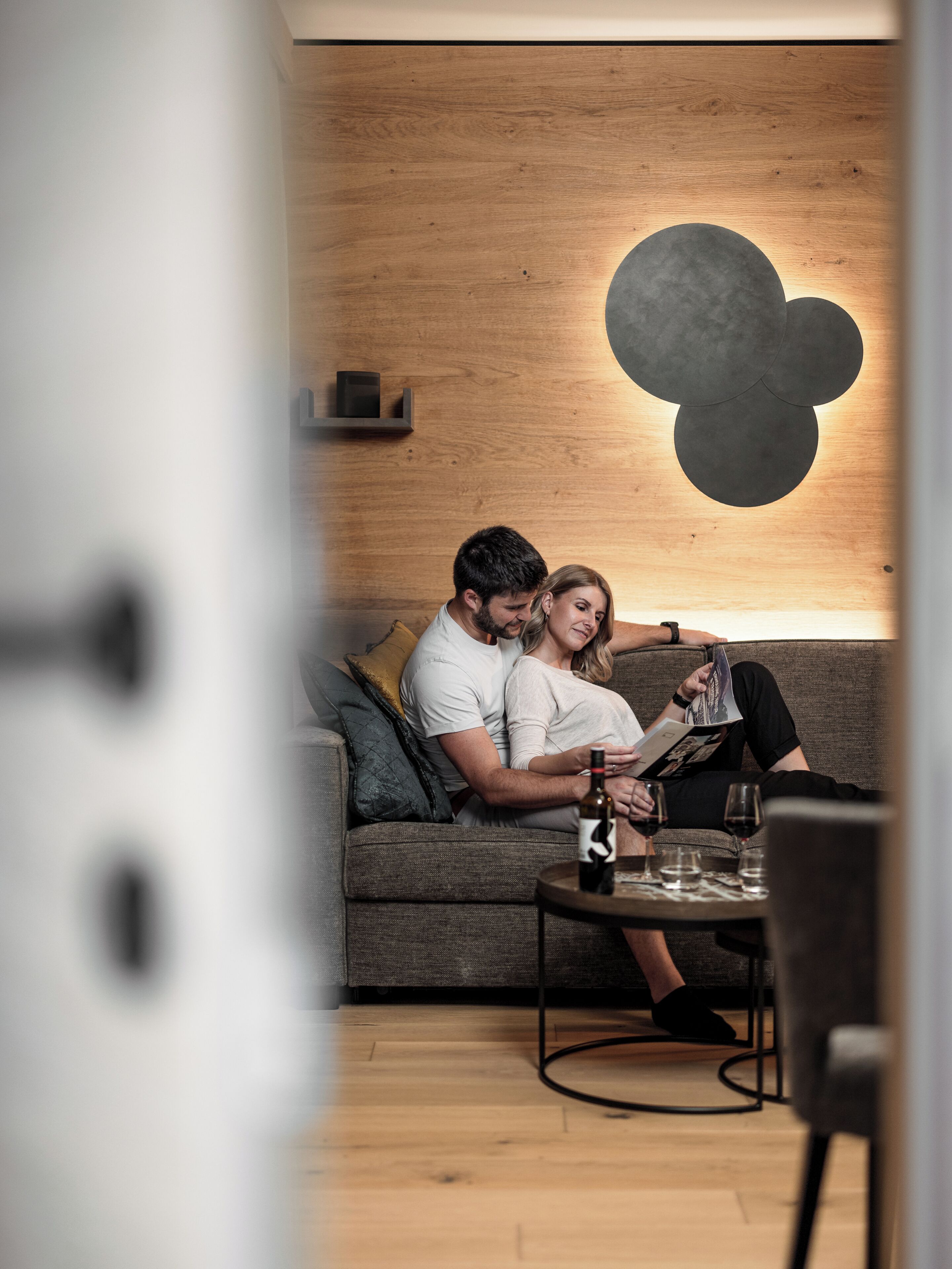A couple on a sofa with a table in front, a wall with a geometric design, and a wooden floor.