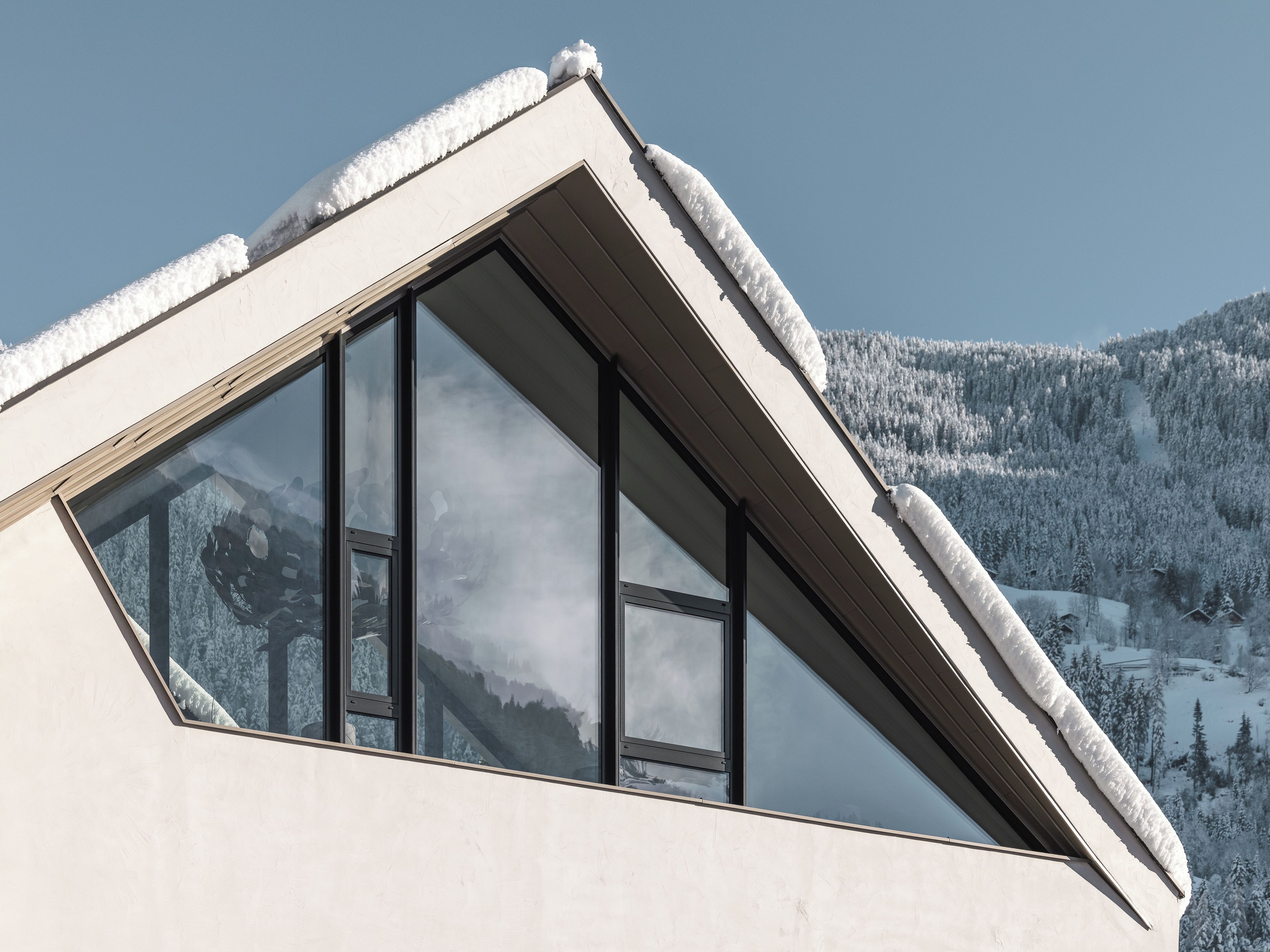 Ein modernes Gebäude mit einem dreieckigen, schneebedeckten Dach, großen Fenstern und Blick auf eine verschneite Berglandschaft.
