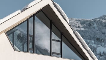 Ein modernes Gebäude mit einem dreieckigen, schneebedeckten Dach, großen Fenstern und Blick auf eine verschneite Berglandschaft.