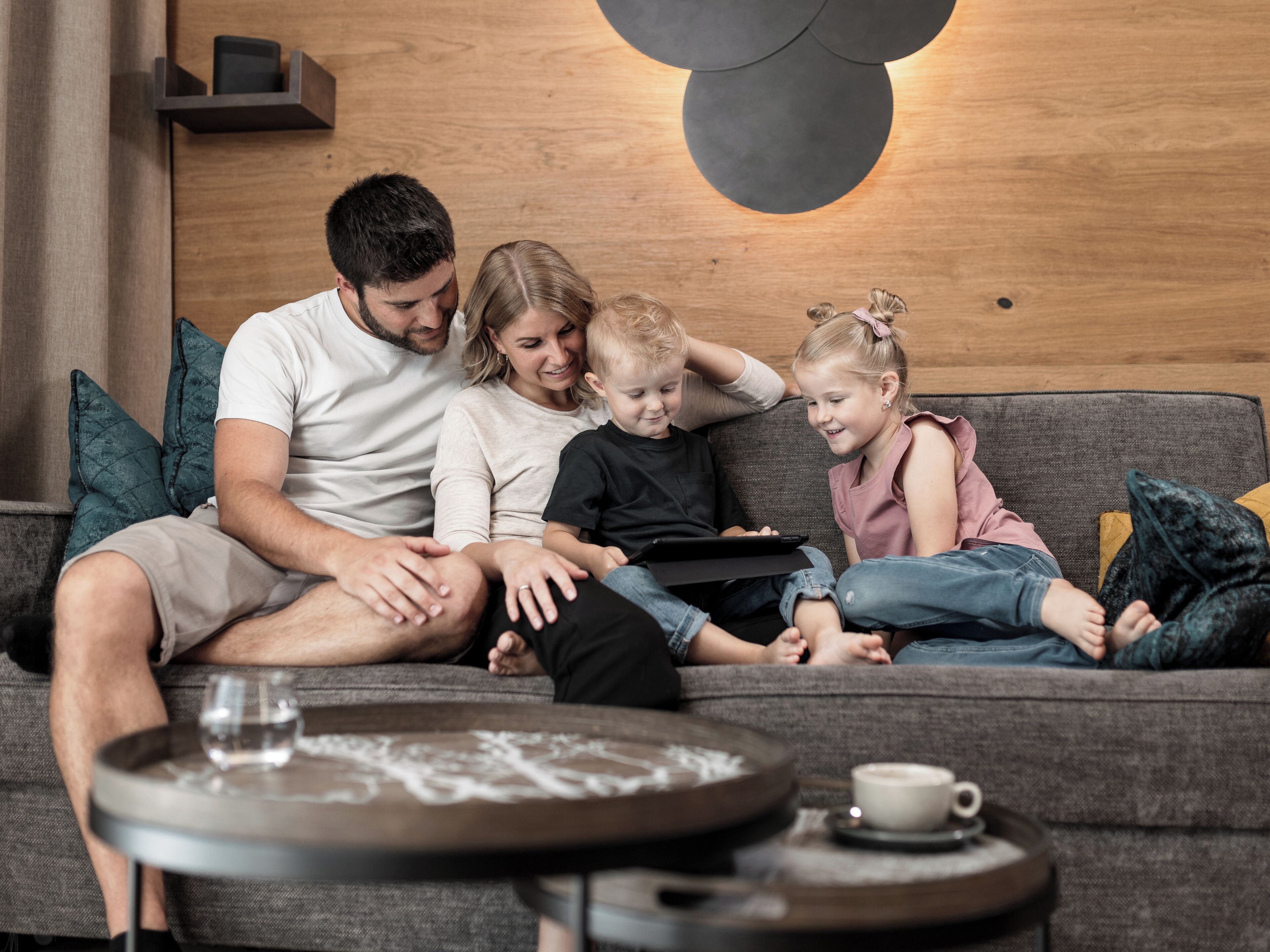 Una familia sentada en un sofá con una tableta, una taza de café sobre una mesita auxiliar, y una pared de madera con una lámpara circular.