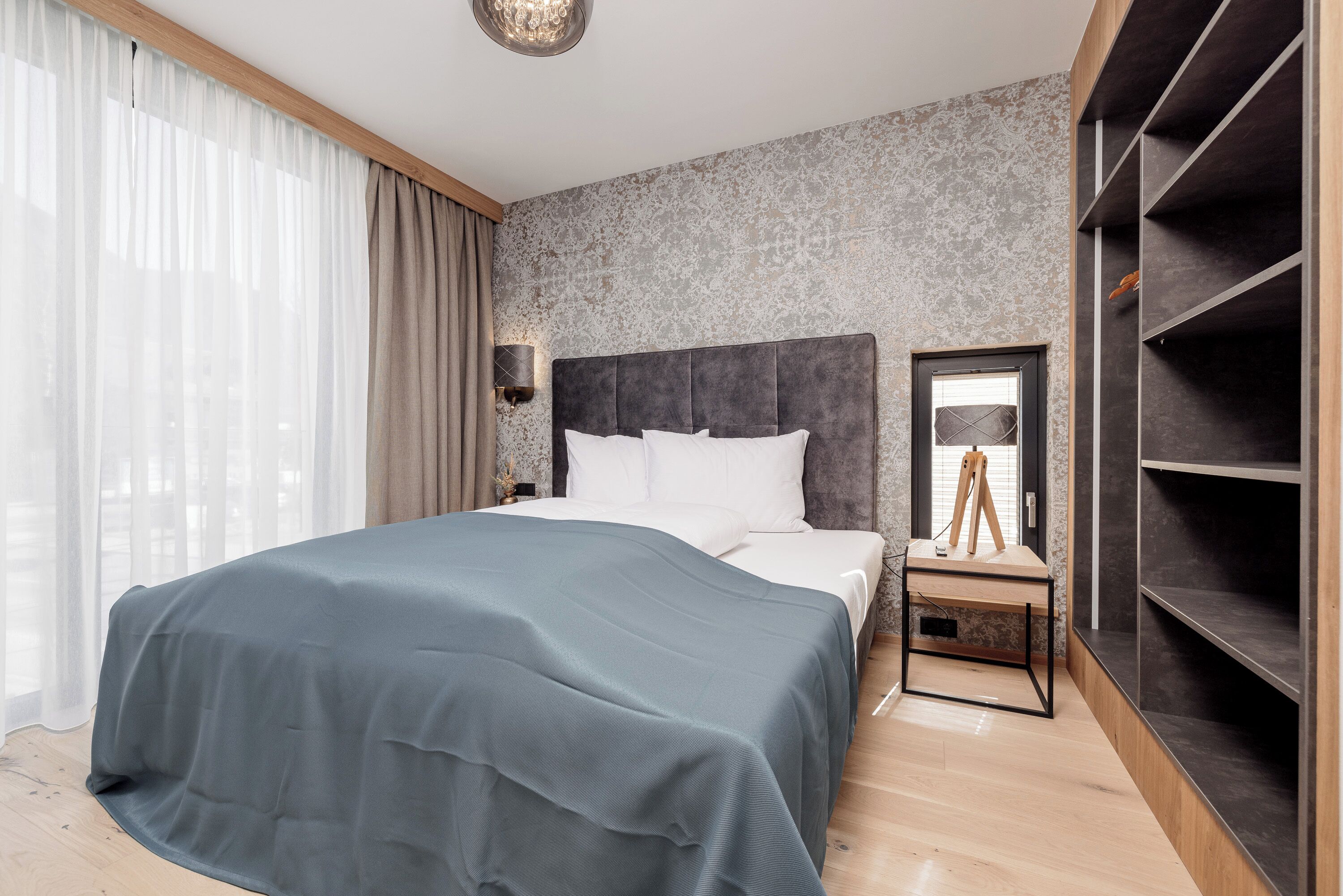 Una habitación de hotel moderna con una cama grande, una mesita de noche con lámpara, un armario y vistas a la ciudad a través de cortinas translúcidas.