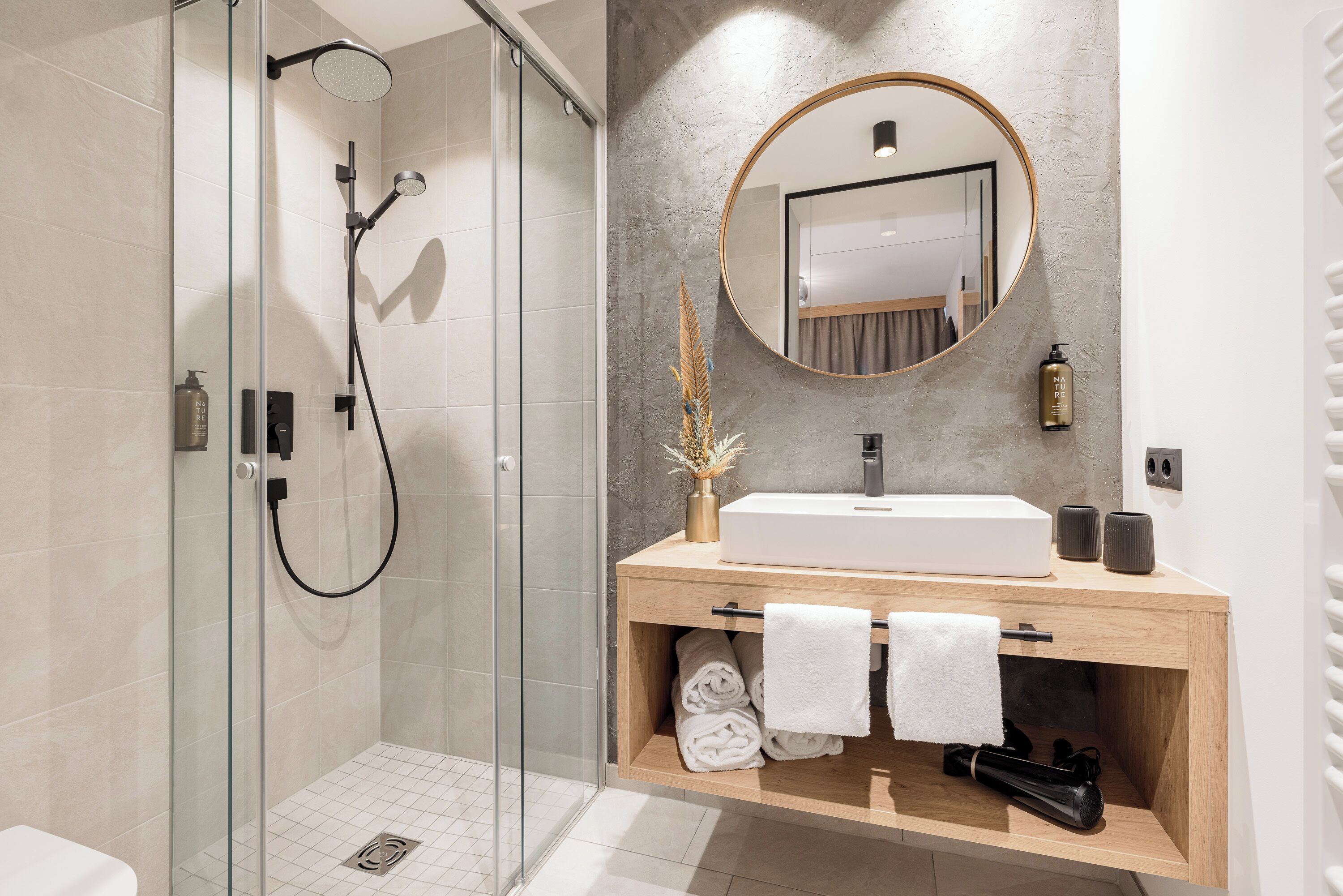 Un baño moderno con ducha envuelta en cristal, tocador de madera con espejo redondo y toallas dobladas con cuidado.