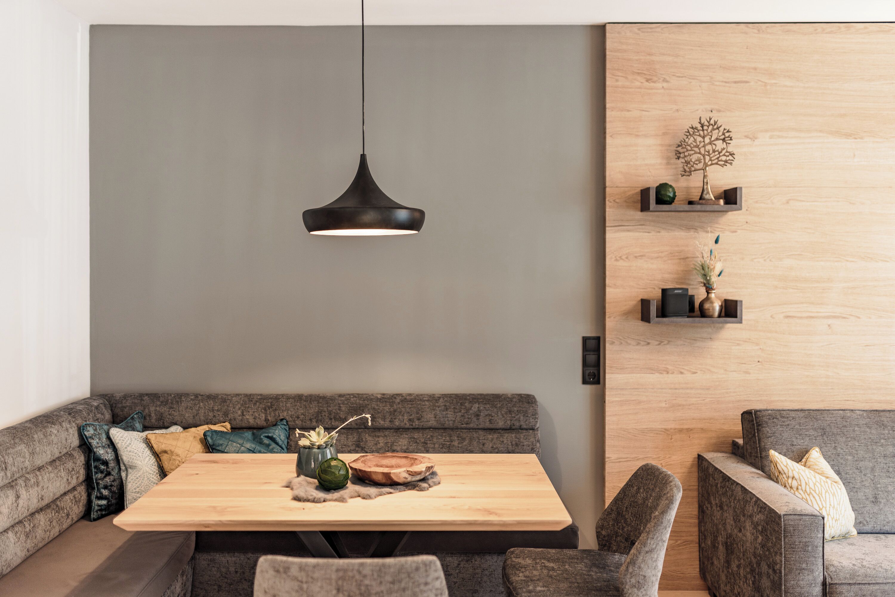 A modern living room with a wooden dining table, grey sofas, and a hanging pendant light.