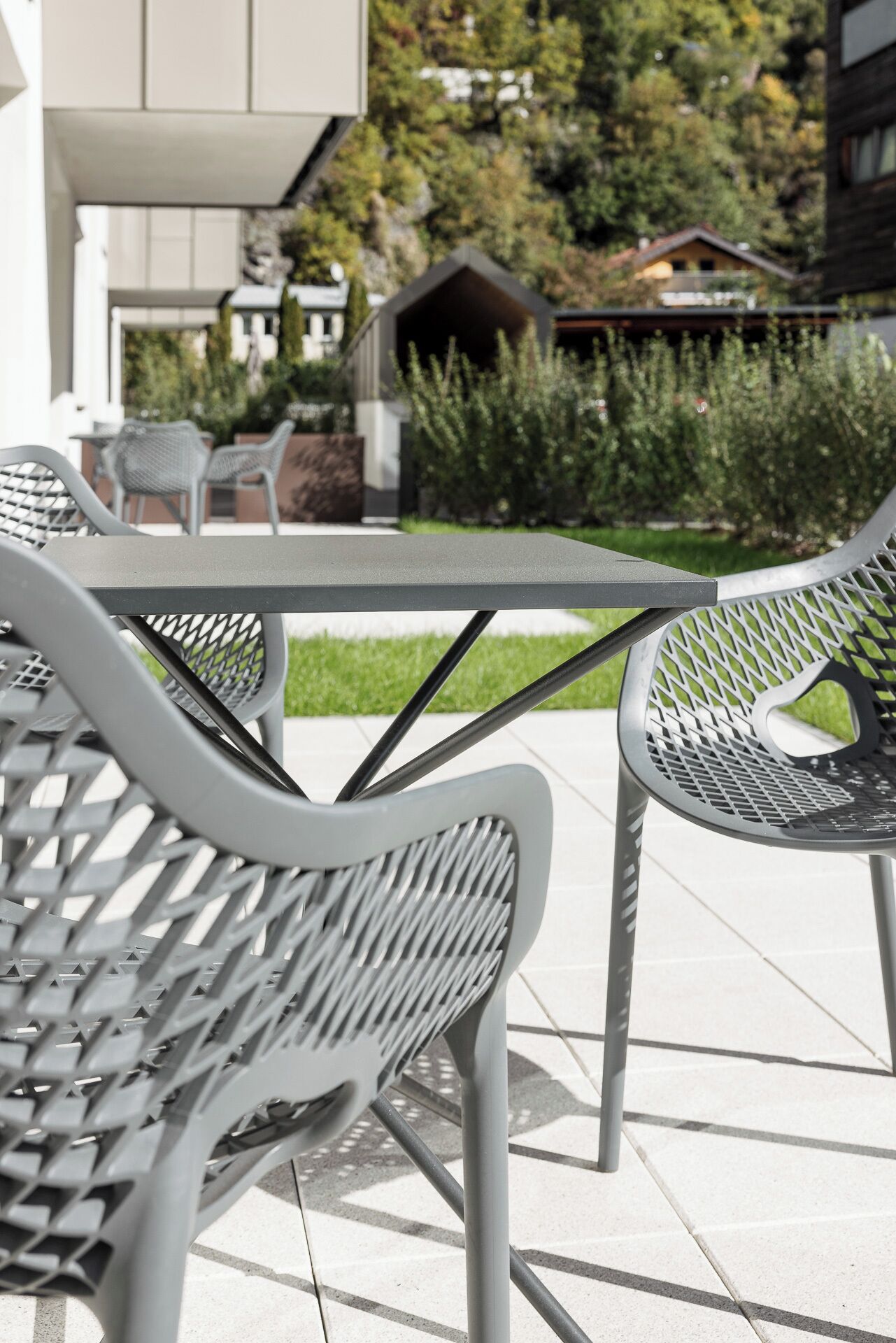 An outdoor seating area with metal chairs and a table, surrounded by a garden and a building.