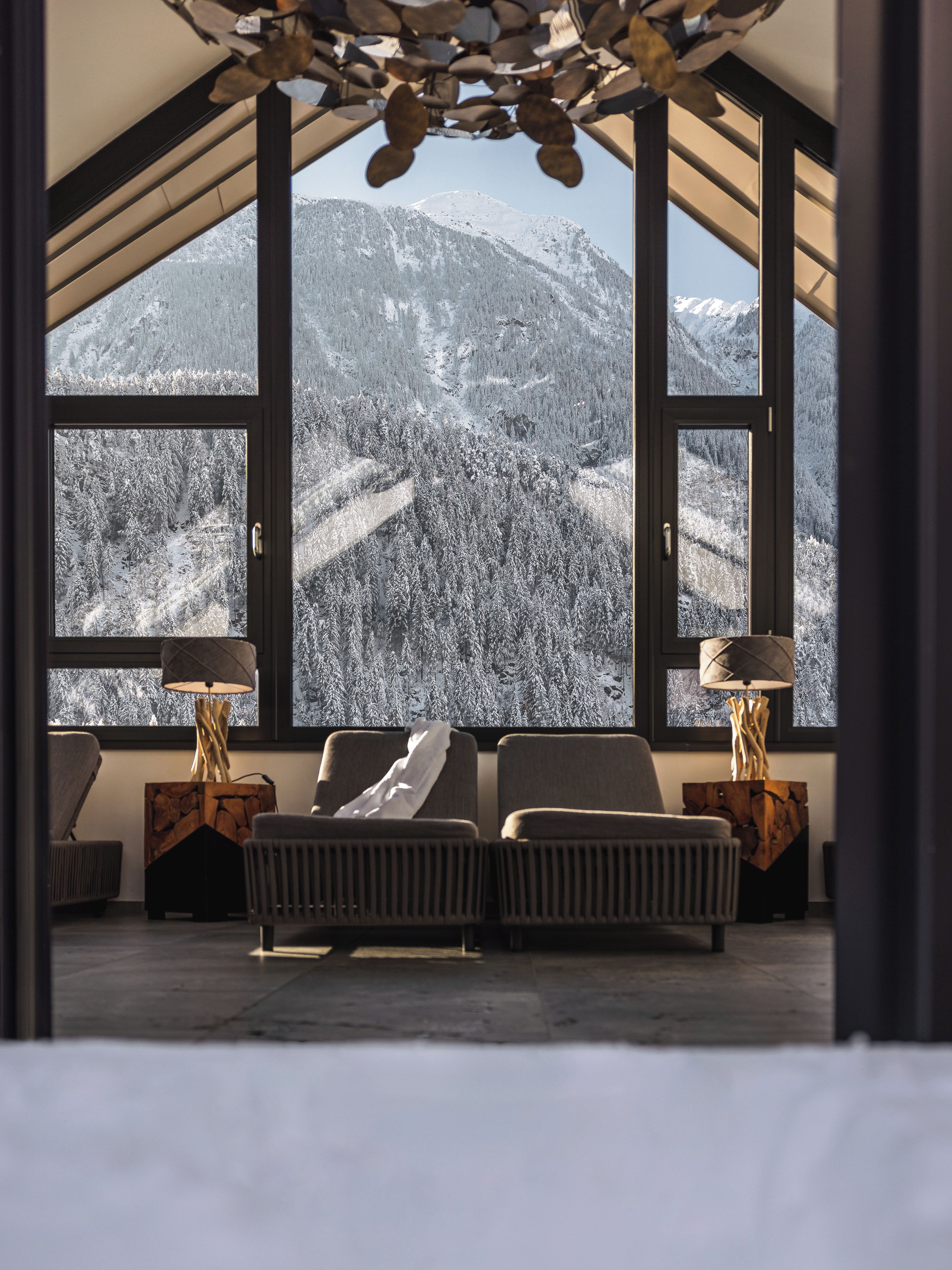 Ein Zimmer mit großem Fenster und Blick auf eine verschneite Berglandschaft, mit zwei Nachttischen inklusive Lampen sowie einer Couch.