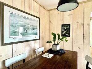 Una habitación con paneles de madera, un cuadro de paisaje marino, una planta en maceta, una mesa con sillas y un cartel que indica Airbnb Pelican Place.