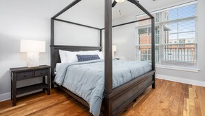 A four-poster bed with a ceiling fan, a nightstand with a lamp, and a window with blinds. - Lavish Apartment over Target in Downtown Auburn - 205 (Auburn)