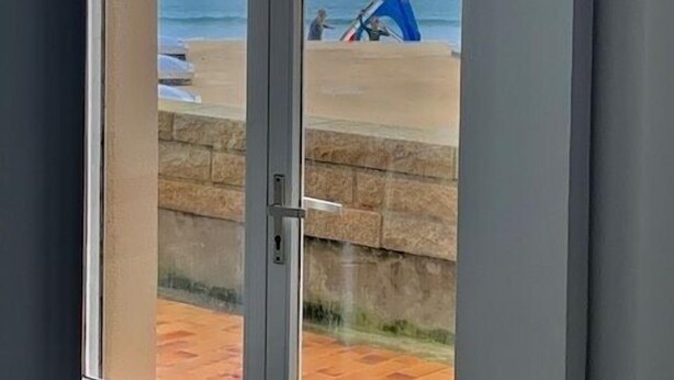 A view through a glass door to a beach with a blue beach chair and a person in the distance.