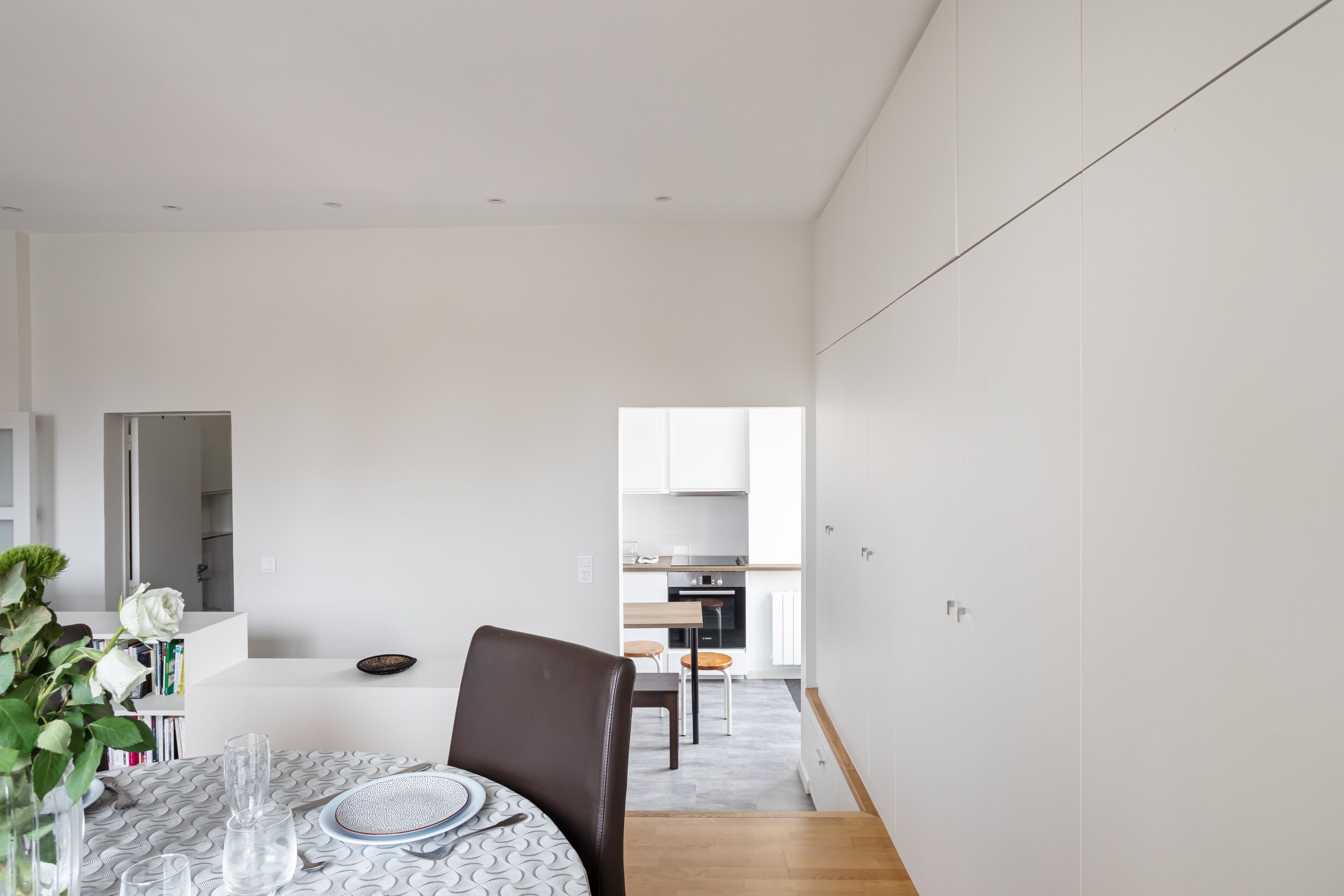 Un espace repas moderne et minimaliste, avec une table dressée pour deux personnes, un vase rempli de fleurs et une vue sur la cuisine.