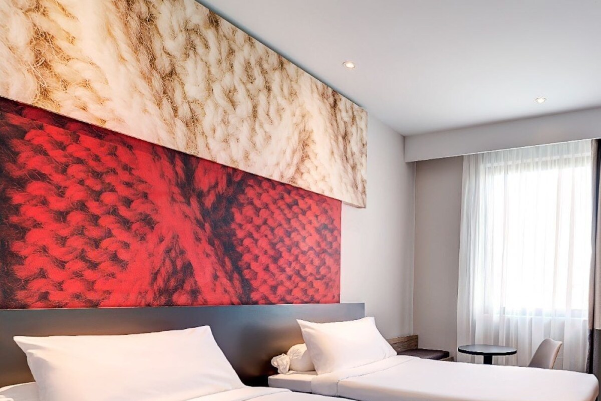 Une chambre d’hôtel avec deux lits, une grande peinture abstraite au mur, une petite table et une fenêtre avec des rideaux.
