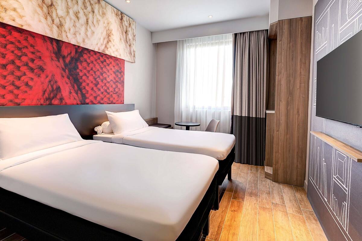 Ein Hotelzimmer mit zwei Betten, einem kleinen Tisch, einem Fernseher und einem großen abstrakten Gemälde an der Wand.