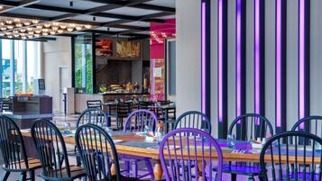 A modern restaurant interior with a long dining table, black and purple chairs, and a kitchen area visible in the background.