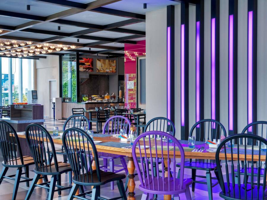 A modern restaurant interior with a long dining table, black and purple chairs, and a kitchen area visible in the background.
