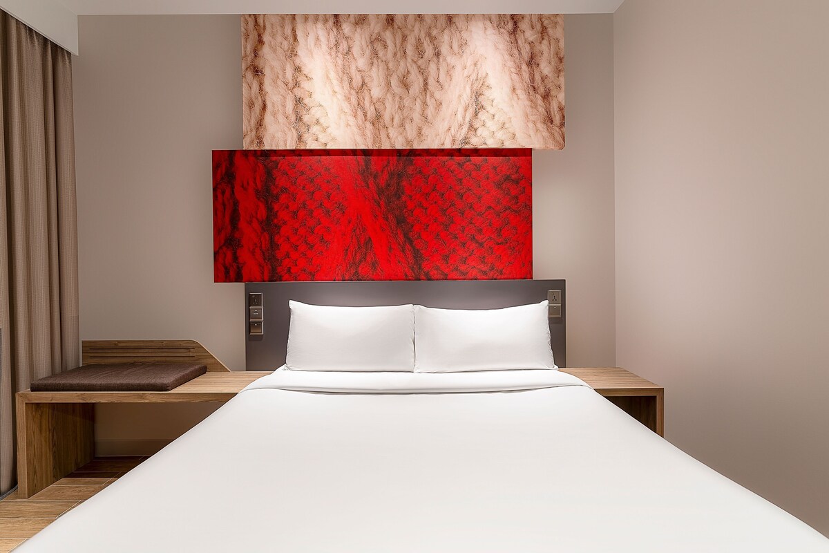 Une chambre moderne avec une tête de lit en bois, une couvre-lit blanche et une œuvre d’art abstraite rouge accrochée au mur au-dessus du lit.