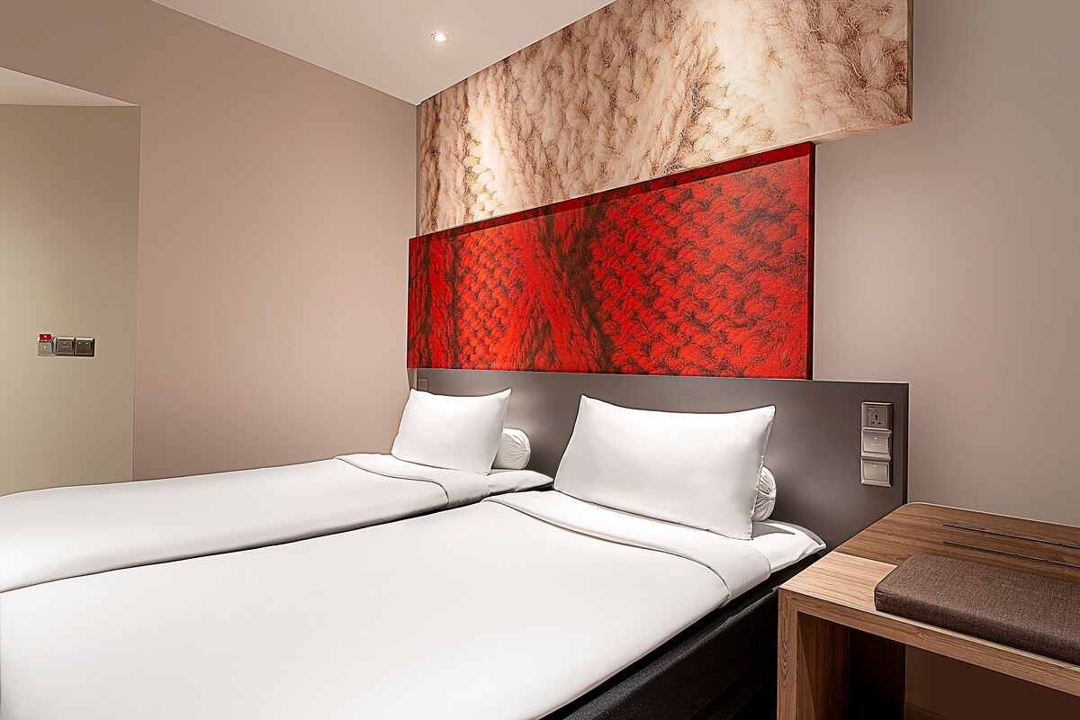 Une chambre d’hôtel moderne avec deux lits, une table de chevet en bois et une œuvre d’art abstraite rouge accrochée au mur.