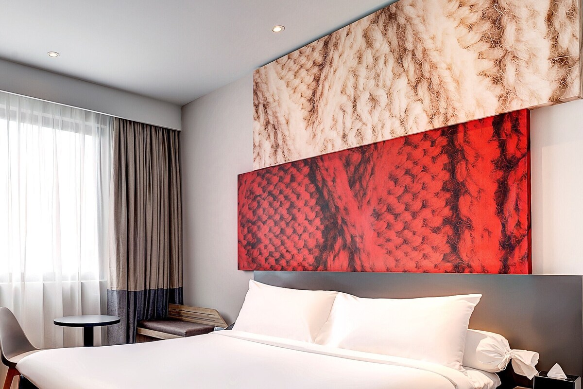 Une chambre d’hôtel moderne avec un grand lit, une petite table ronde, une chaise et une table de chevet avec une lampe.