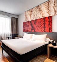 Una habitación de hotel moderna con una cama grande, una mesita redonda pequeña, una silla y una mesita de noche con lámpara.