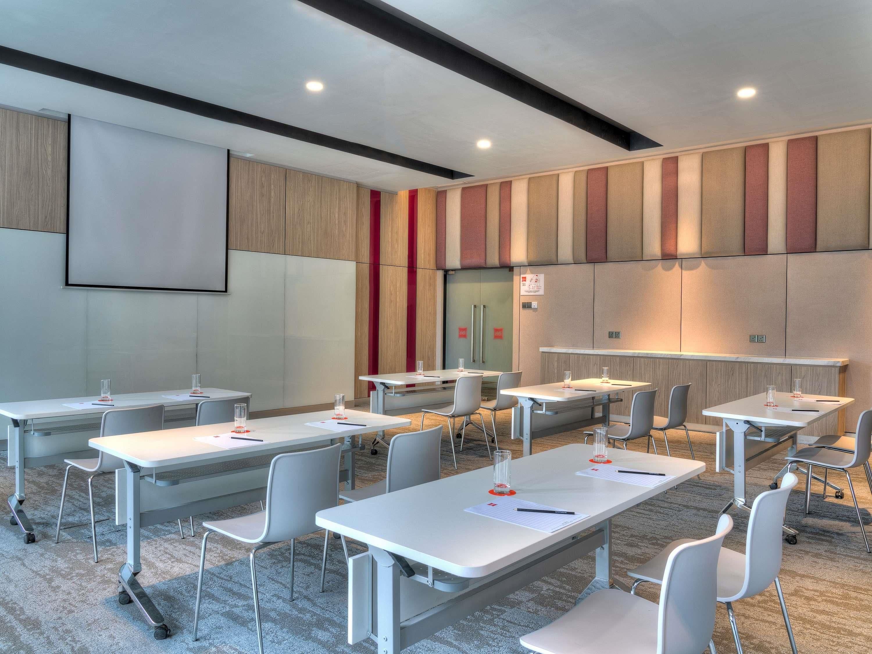 A conference room with white tables, grey chairs, a projector screen, and a red accent wall.