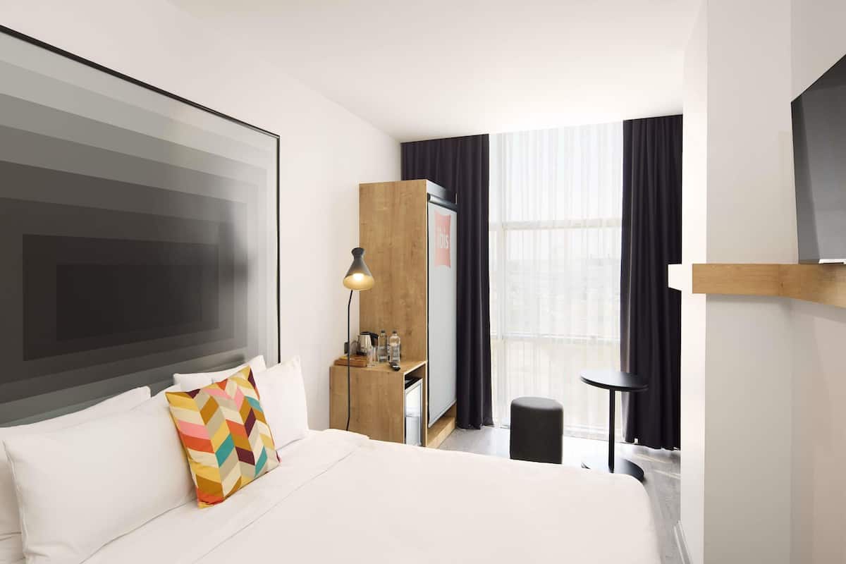 A hotel room with a large bed, a desk, a chair, a TV, and a window with curtains.