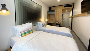 Une chambre d’hôtel moderne avec deux lits simples, une grande peinture abstraite, un petit bureau avec une lampe et une porte donnant sur un couloir.