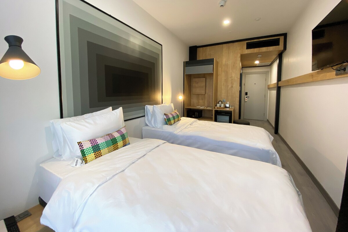 A modern hotel room with two single beds, a large abstract painting, a small desk with a lamp, and a door leading to a hallway.