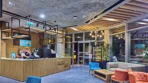 A modern cafe interior with a wooden reception desk, seating area with sofas and chairs, and large windows allowing natural light. - Ibis Istanbul Airport (Arnavutköy)