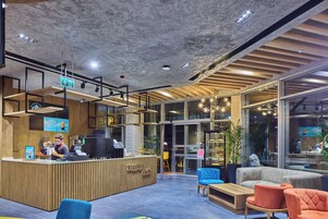 Interior de una cafetería moderna con un mostrador de recepción de madera, zona de estar con sofás y sillones, y grandes ventanales que dejan entrar luz natural.