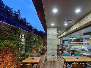 An outdoor café with wooden tables and chairs, a brick wall with climbing plants, and a glass partition separating the dining area from the kitchen. - Ibis Istanbul Airport (Arnavutköy)