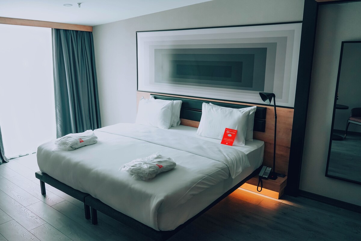 A modern hotel room with a neatly made bed, a large window with curtains, a flat-screen TV mounted on the wall, and a bedside table with a lamp.