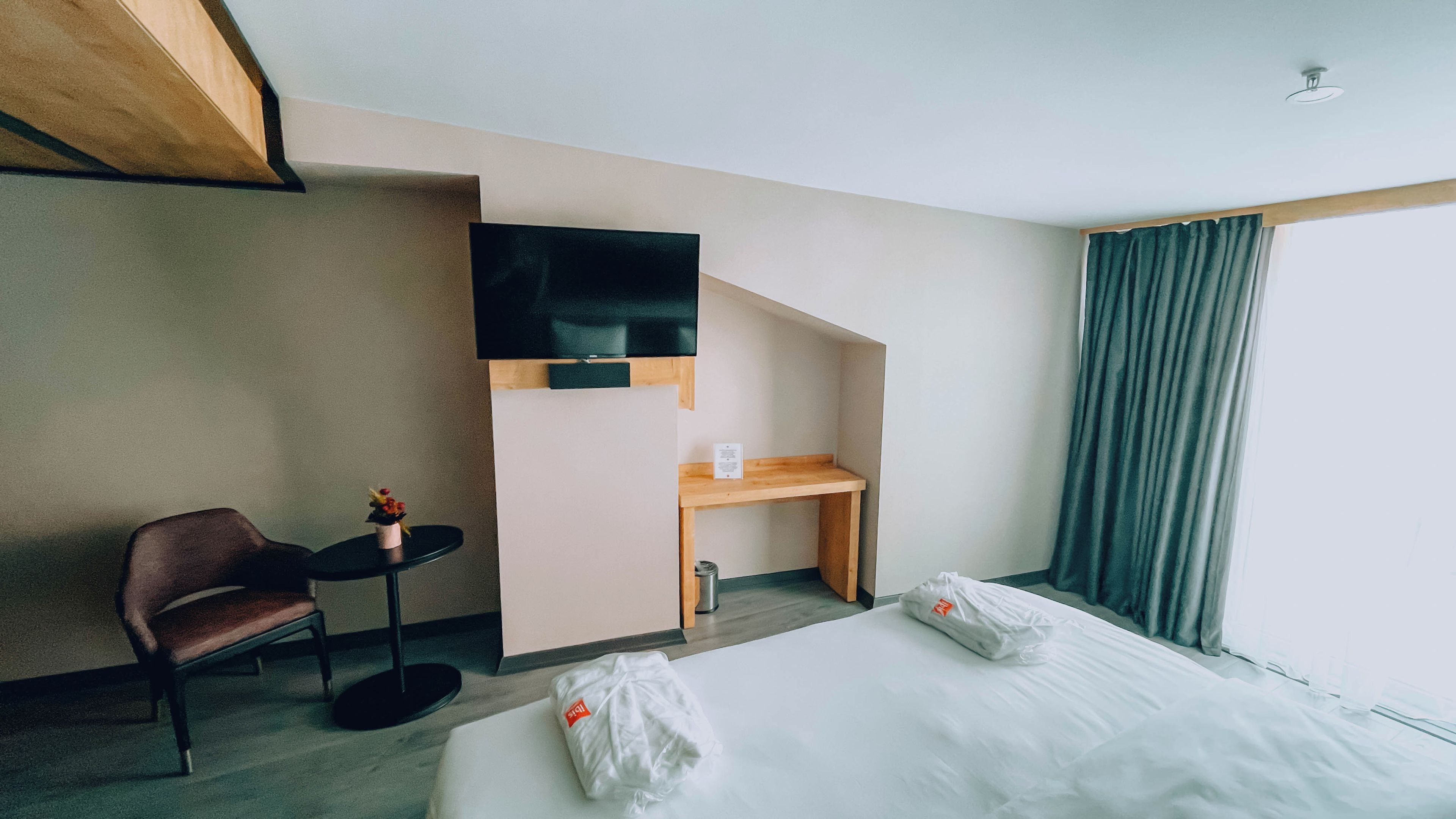 Habitación de hotel con cama, escritorio, silla, televisión y ventana con cortinas.