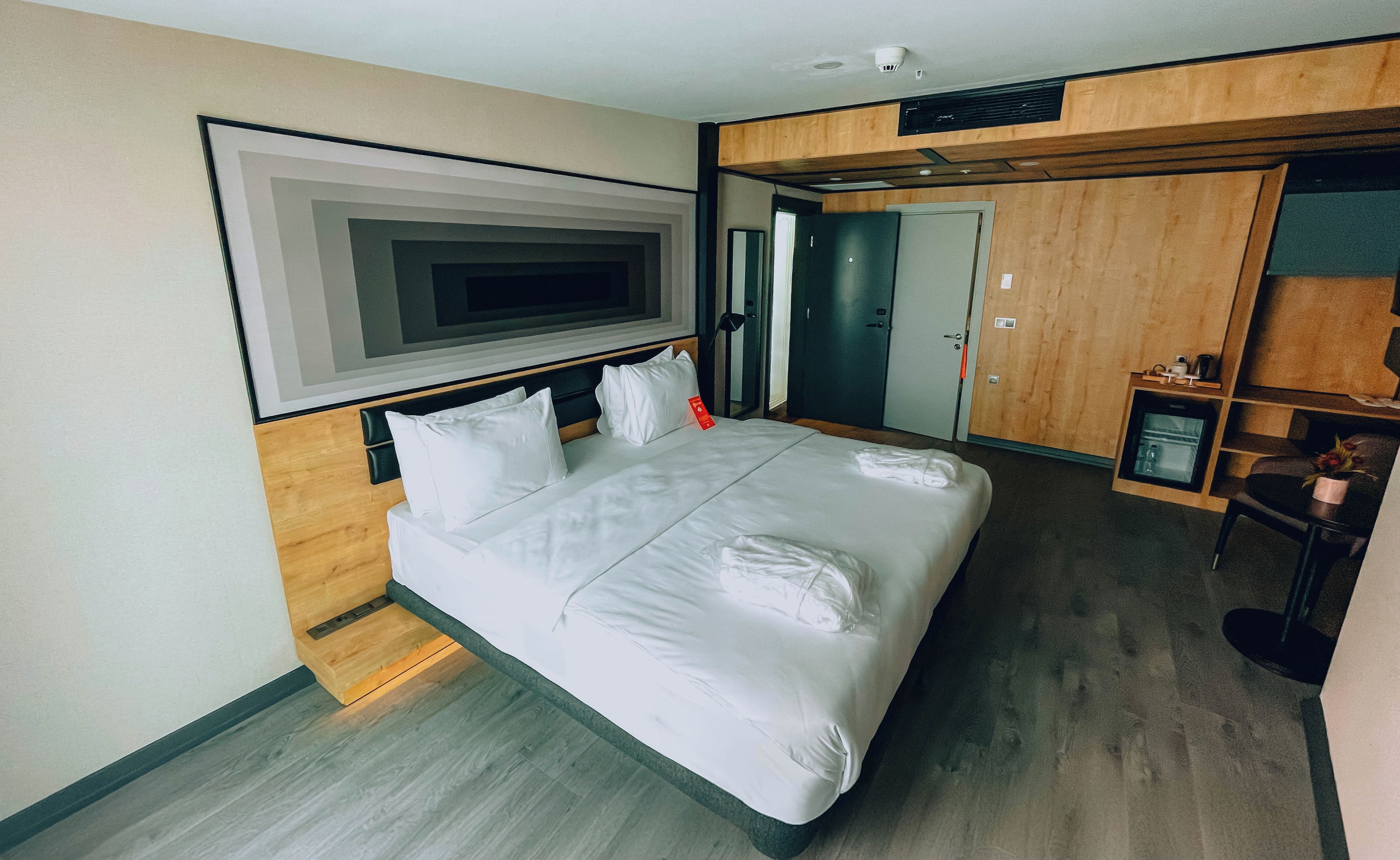 A hotel room with a large bed, a television mounted on the wall, a wardrobe, and a desk with a chair.