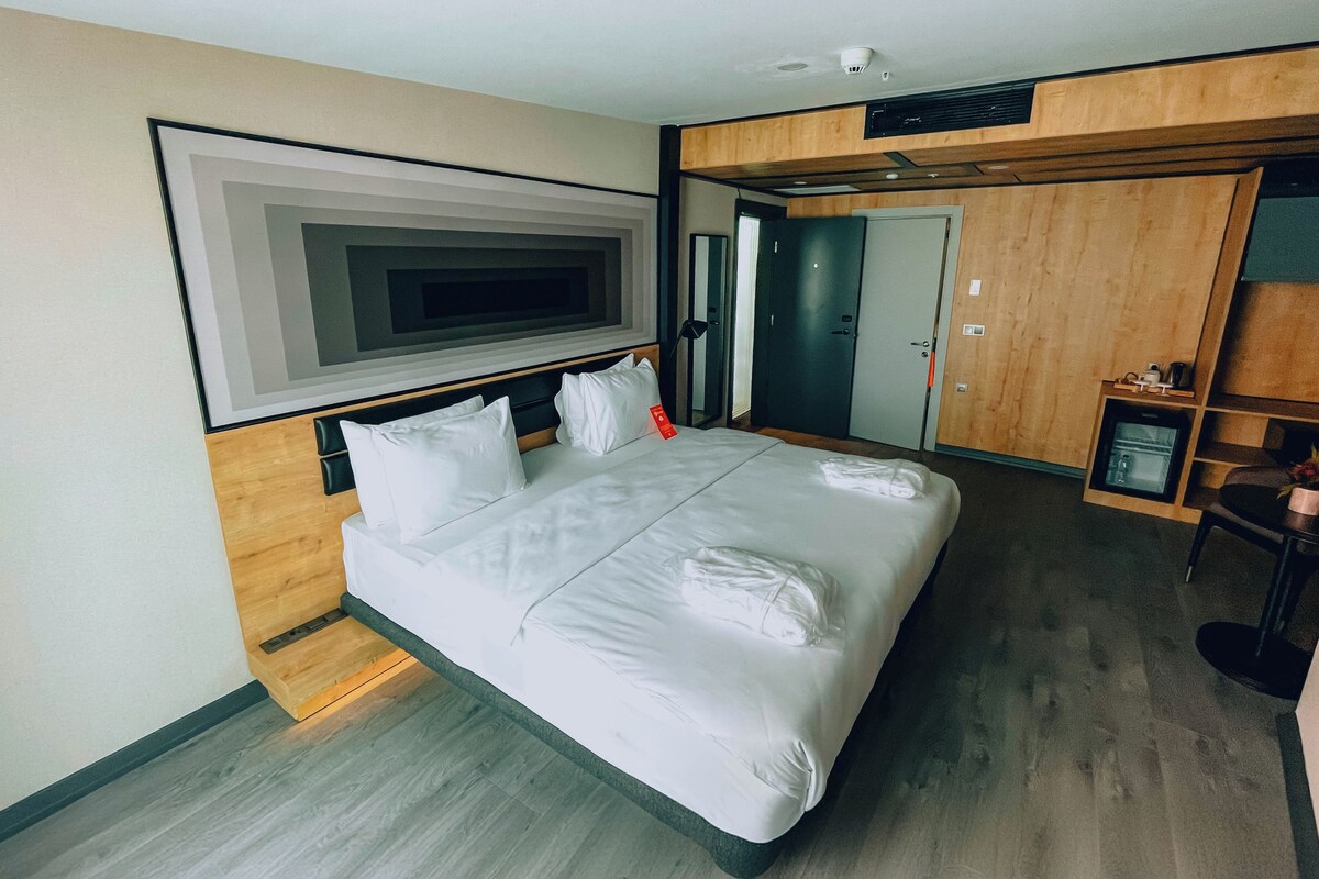 A hotel room with a large bed, a flat-screen TV mounted on the wall, a wooden wardrobe, and a small seating area with a chair and table.