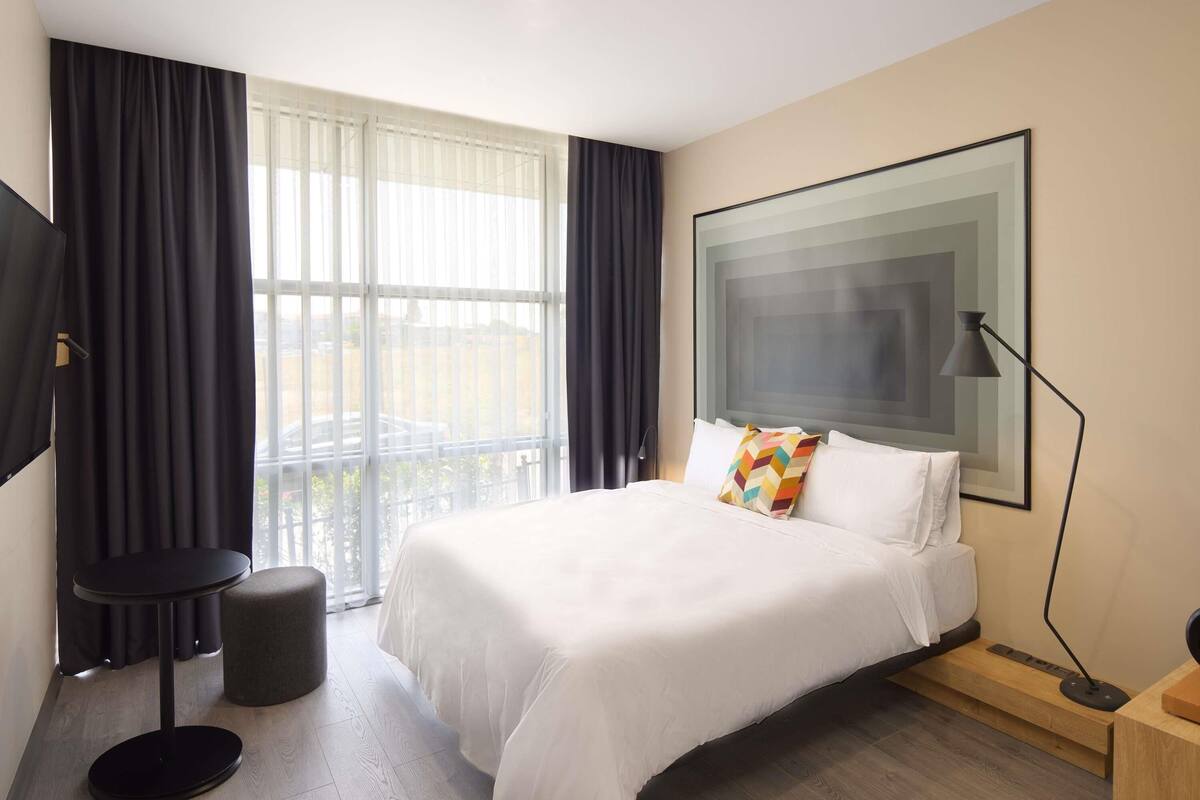 A modern hotel room with a large bed, a flat-screen TV, a desk with a lamp, and a window with sheer curtains.