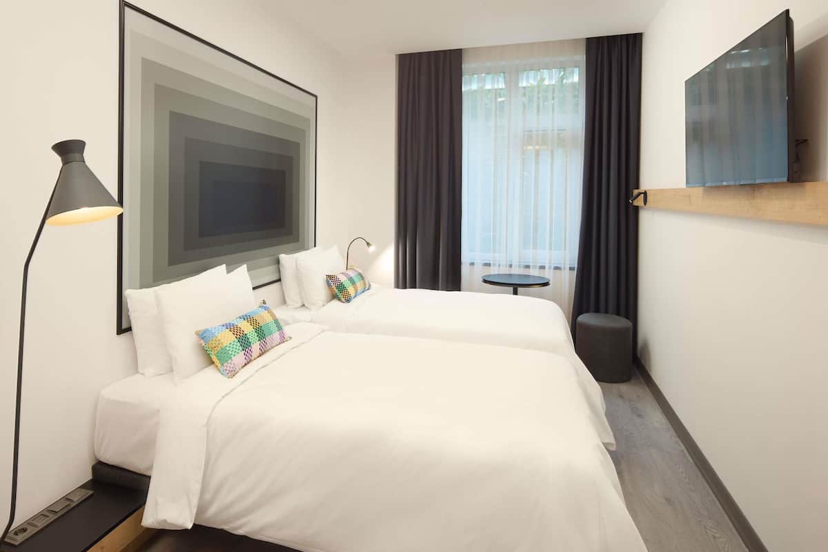 A hotel room with a bed, a desk, a chair, a lamp, a TV, a window with curtains, and a view of the outdoors.