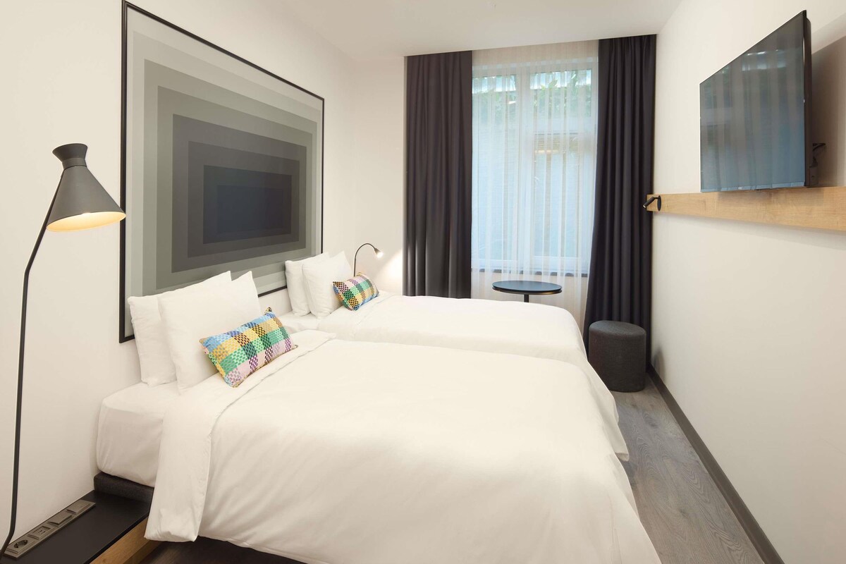 A hotel room with a bed, a desk, a chair, a lamp, a TV, a window with curtains, and a view of the outdoors.