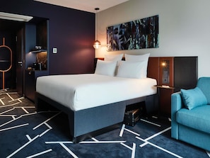 Une chambre d’hôtel moderne avec un lit, un fauteuil bleu, un tableau au mur et un tapis à motifs géométriques.