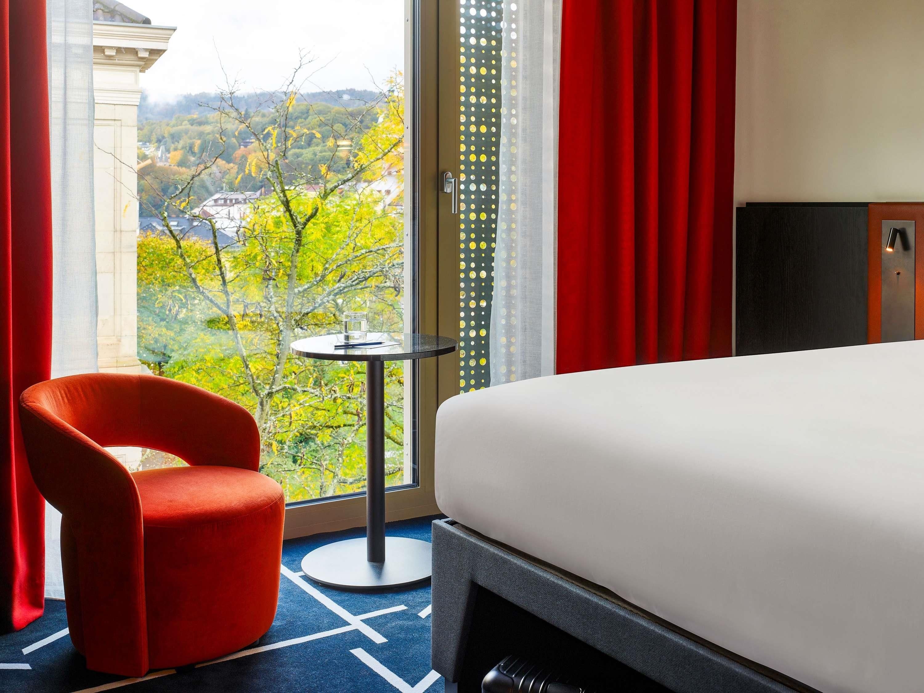 Ein Hotelzimmer mit Bett, einem roten Sessel, einem kleinen Tisch und Blick auf eine grüne Landschaft.