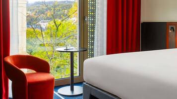 Ein Hotelzimmer mit Bett, einem roten Sessel, einem kleinen Tisch und Blick auf eine grüne Landschaft.