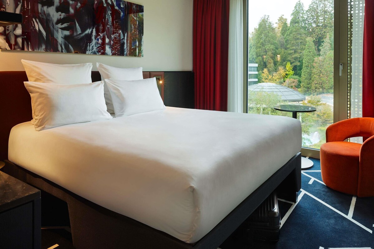 A hotel room with a large bed, a desk, a chair, a painting on the wall, and a view of trees through the window.