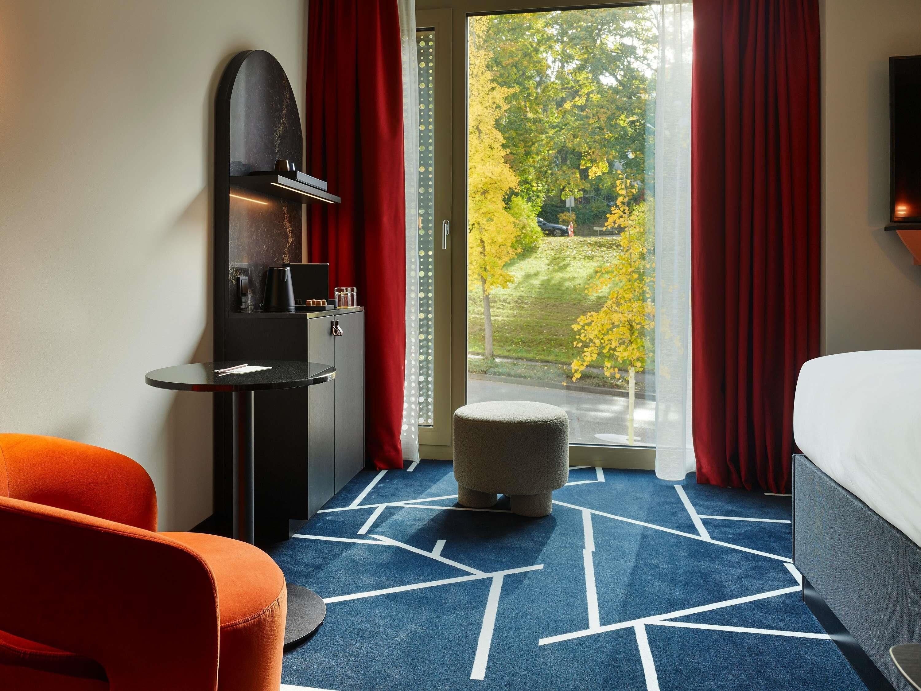 Ein Hotelzimmer mit Bett, Schreibtisch mit Kaffeemaschine, Stuhl und Blick auf Bäume durch ein Fenster.