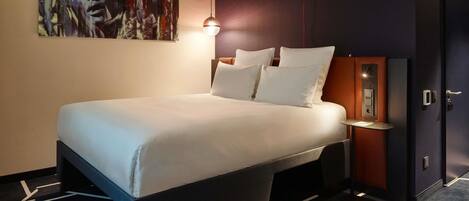 A hotel room with a bed, a nightstand, a wall-mounted light, a door, and a painting on the wall.