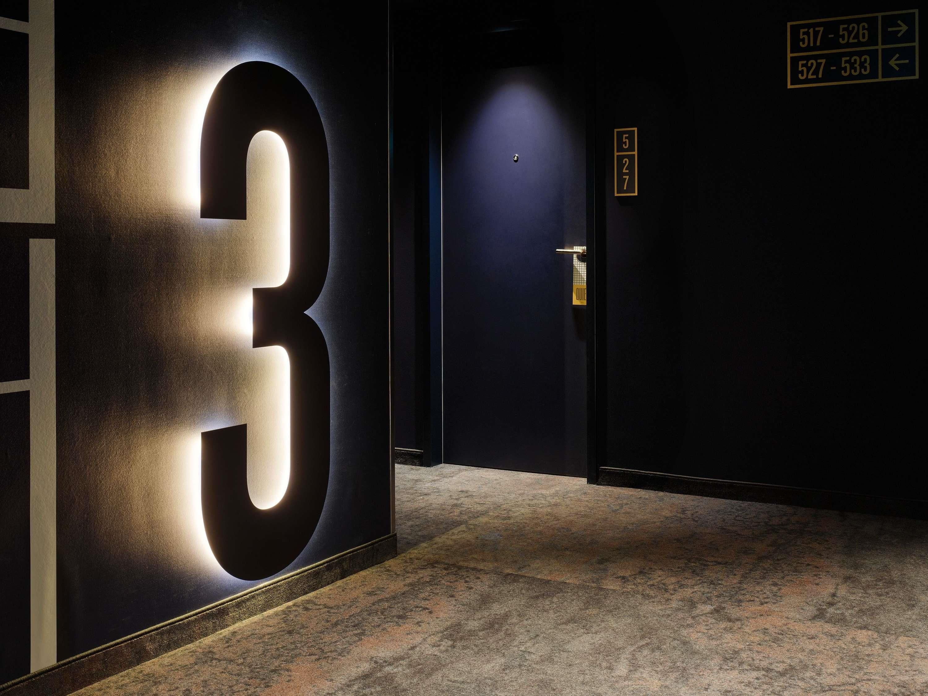 Un pasillo con un letrero grande e iluminado con el número '3', una puerta con cerradura electrónica y señalización que indica los números de habitación.