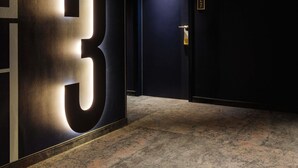 Un couloir avec un grand panneau lumineux affichant le chiffre « 3 », une porte dotée d’un clavier pour l’ouverture à code, et des panneaux indicateurs indiquant les numéros des pièces.