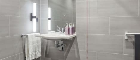 Un baño moderno con un lavamanos blanco, dos grifos y un espejo. Hay artículos de aseo en el lavamanos y una toalla colgando en un perchero.