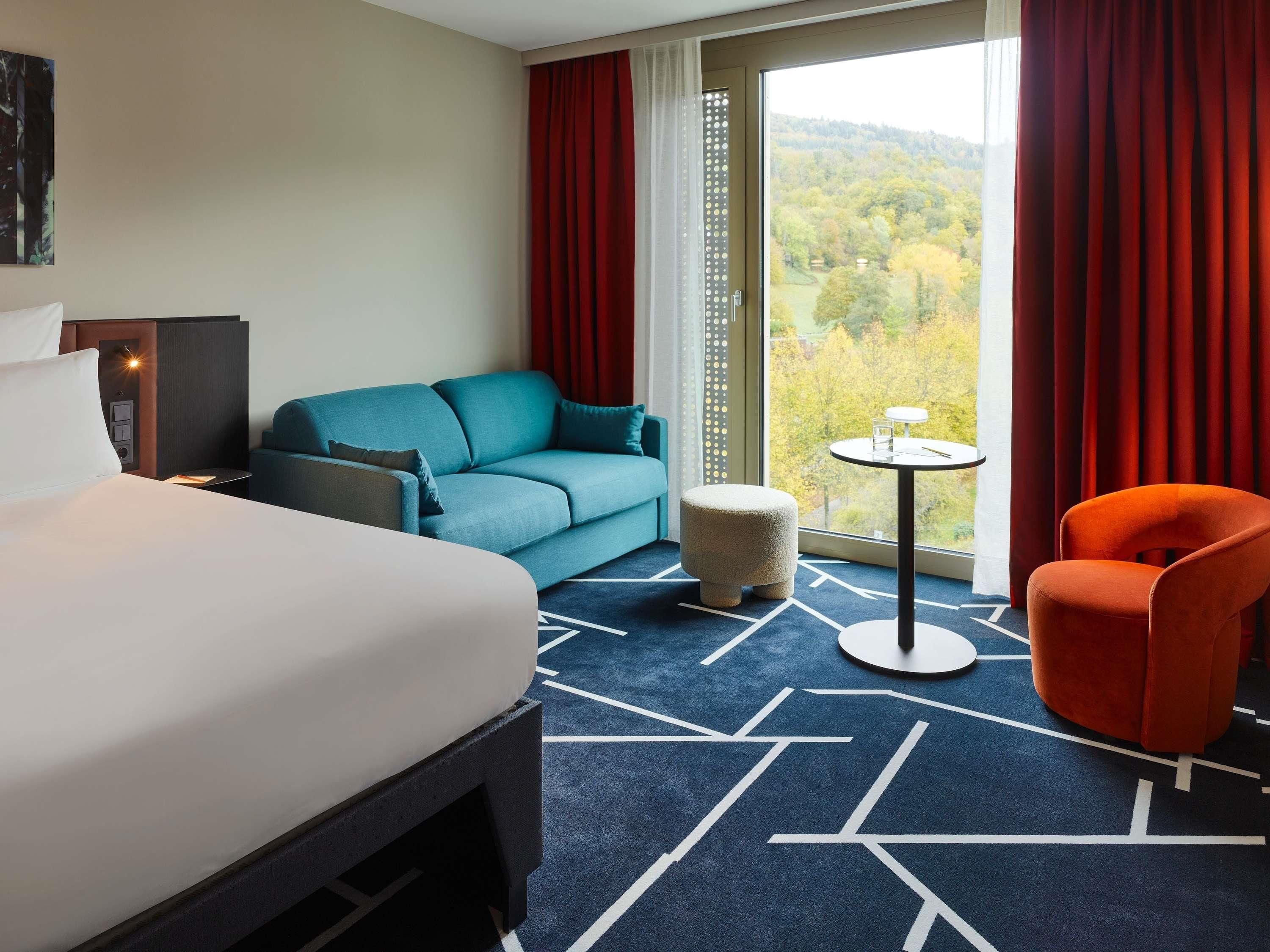 Una habitación de hotel con una cama, un sofá, una mesita redonda, una silla y vistas a árboles a través de una ventana.